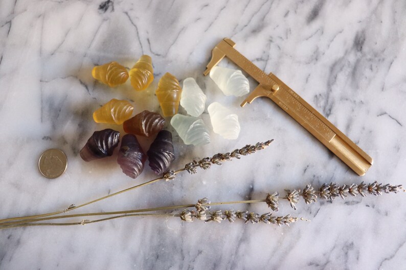 May include: A collection of colorful, translucent, shell-shaped candies on a marble surface. The candies are arranged in a cluster, with some overlapping. A gold-colored caliper is placed to the right of the candies, and a dried lavender sprig is placed below the candies.