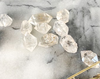 Könnte beinhalten: Eine Sammlung von klaren Quarzkristallen, auch bekannt als Herkimer-Diamanten, mit einer facettierten, doppelt abgeschlossenen Form. Die Kristalle sind auf einer weißen Marmorfläche angeordnet.
