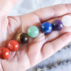 May include: A hand holding seven smooth, round, polished stones in a rainbow of colours, including red, orange, yellow, green, blue, purple, and white.