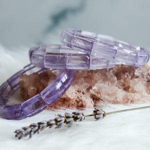 Puede incluir: Dos pulseras translúcidas de color morado claro compuestas por cuentas rectangulares de piedra pulida. Las pulseras están apiladas y descansan sobre un racimo mineral rugoso de color marrón rosáceo claro, junto a una rama de lavanda seca.