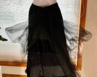 May include: Black tiered skirt with a flowing tulle overlay. The skirt features multiple layers of fabric, including velvet and satin, with lace trim. The tulle adds volume and a dramatic flair to the garment. The skirt is displayed on a mannequin.