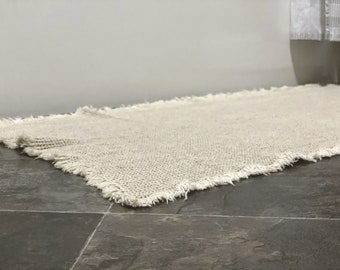 May include: An off-white rectangular rug with a fringed edge, made from a coarse, woven material. The rug sits on a dark tiled floor. The natural colour and texture of the rug suggest a rustic or minimalist design, suitable for a bathroom or hallway.