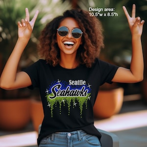 Peut inclure: T-shirt noir avec un motif en strass. Le motif présente le texte "Seattle Seahawks" en bleu et vert, avec un effet scintillant. La zone de conception est de 26,7 cm de large x 21,6 cm de haut.