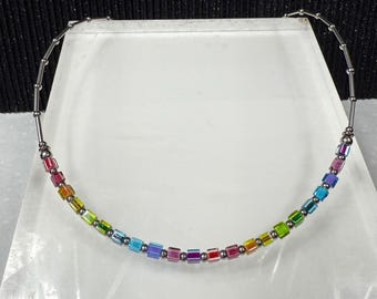 May include: A rainbow-colored beaded necklace with a silver-tone metal chain. The necklace features small, square beads in a spectrum of colors, including red, orange, yellow, green, blue, and purple. The beads are strung together with small silver-tone metal links.