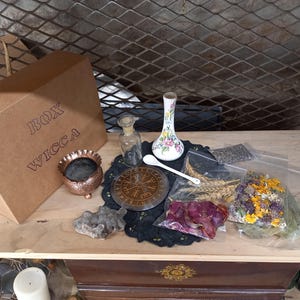 Op de afbeelding: Een verzameling Wicca-artikelen op een houten oppervlak. Inclusief een kartonnen doos met "BOX WICCA" erop gedrukt, een koperen wierookbrander, een kleine glazen fles, een bloemenvaas, een witte lepel, een kompas en gedroogde kruiden en bloemen.