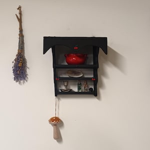 May include: A black wooden shelf with three shelves, decorated with red cherries. The top shelf has a red ceramic pot, the middle shelf has a white plate, and the bottom shelf has a small candle holder, three small glass bottles, and a small black object. A brown mushroom ornament hangs from the bottom shelf.