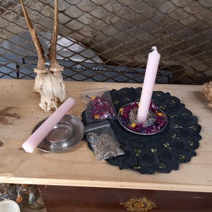 May include: A selection of spiritual items on a wooden surface. Includes a pink candle, a metal dish, dried flowers, and a deer skull. A black lace doily and a decorative rock are also present.