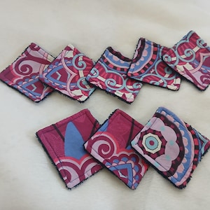 Può includere: Un set di otto sottobicchieri quadrati in tessuto. Ogni sottobicchiere presenta un motivo ripetuto in tonalità di rosa, viola, blu e bianco. I bordi sono rifiniti con un bordo scuro e strutturato, creando un oggetto decorativo e funzionale.