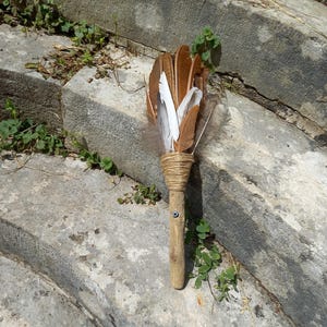Può includere: Una bacchetta di piume fatta a mano con un manico di legno. La bacchetta presenta un fascio di piume marroni e bianche, legate con spago. Una piccola perla con occhio blu è inserita nel manico di legno. La bacchetta è su una superficie di pietra usurata.