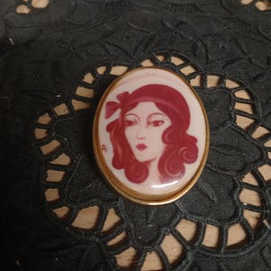 Peut inclure: Broche ovale vintage avec un portrait de femme. Le visage de la femme est peint en rouge et blanc, avec un chapeau rouge et des cheveux bouclés. La broche est encadrée d'un métal doré.