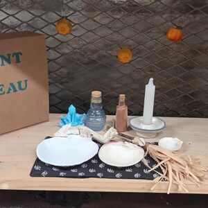 Può includere: Una scatola di cartone marrone con il testo "ELEMENT EAU" sul davanti. La scatola è appoggiata su uno scaffale di legno con una varietà di oggetti, tra cui una candela bianca, due bottiglie di vetro, una piccola ciotola e una conchiglia.