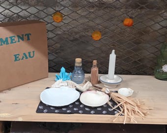 May include: A brown cardboard box with the text "ELEMENT EAU" on the front. The box is sitting on a wooden shelf with a variety of items, including a white candle, two glass bottles, a small bowl, and a seashell.