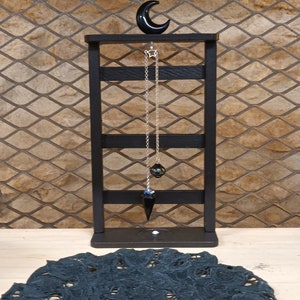 Peut inclure: Un collier pendule noir avec un charme en forme d'étoile et de lune, exposé sur un présentoir en bois noir. Le présentoir est posé sur une surface en bois, avec un napperon en dentelle noire et une machine à coudre au premier plan.