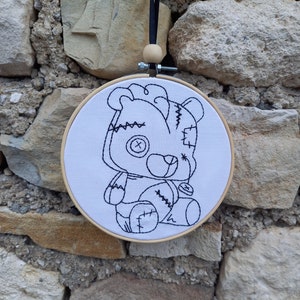 Peut inclure: Un cercle à broder en bois avec un tissu blanc orné d'un motif cousu en noir représentant un ours en peluche. L'ours a des patchs, un œil bouton et une bouche cousue. Un ruban noir et une perle en bois sont attachés pour l'accrocher.