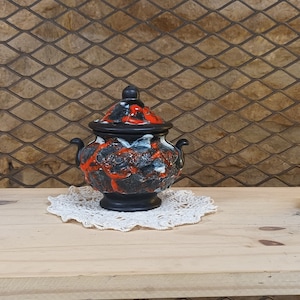 Può includere: Un vaso in ceramica nero e arancione con un coperchio, seduto su un pizzo bianco su una superficie di legno. Il vaso ha un design floreale bianco e arancione.