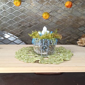 Op de afbeelding: Een glazen pot met een blauw-wit deksel gevuld met gedroogde kruiden en kristallen. De pot staat op een groene gehaakte onderzetter. Er hangen kleine oranje pompoenen aan een draadgaas achtergrond.