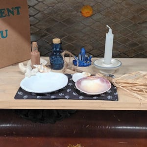 Può includere: Un piccolo scaffale di legno con una scatola etichettata "ELEMENT EAU" e una varietà di oggetti, tra cui una bottiglia di vetro blu, un piatto bianco, una conchiglia, una candela e un fascio di paglia.