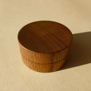 May include: A small, round wooden box with a dark brown finish. The lid is closed and the box is sitting on a light brown surface.