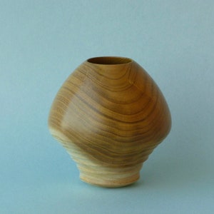 May include: A wooden vase with a light brown colour and a swirling grain pattern. The vase has a rounded, bulbous shape with a slightly flared opening.