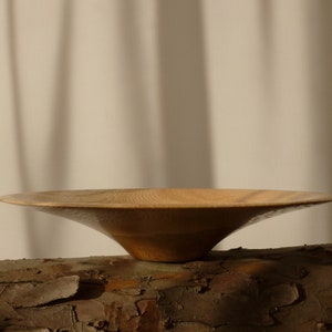 May include: A wooden bowl with a wide, shallow base and a gently sloping, rounded interior. The bowl is made of light brown wood with a natural grain pattern. It is resting on a piece of tree bark.