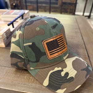 May include: Camouflage baseball cap with a brown leather patch featuring a black and white American flag. The cap has a curved brim and a button on top. The camouflage pattern includes shades of green, brown, and beige.