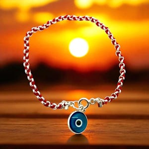 May include: A red and silver bracelet with a blue eye charm. The bracelet features a woven red and silver chain design. The charm is a round, blue eye with a black center, attached with a silver clasp. The background is a sunset.