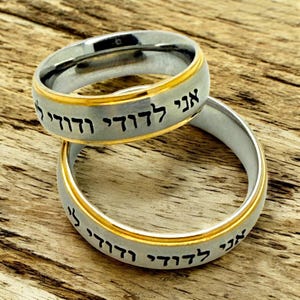 May include: Two silver-colored rings with a gold-colored band and Hebrew text. The rings are stacked on top of each other, with the text visible on the outer band. The rings are set against a wooden background.