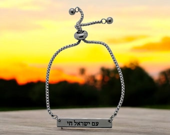 Puede incluir: Un brazalete ajustable de plata con un dije de barra de plata que dice "עם ישראל חי" en hebreo.