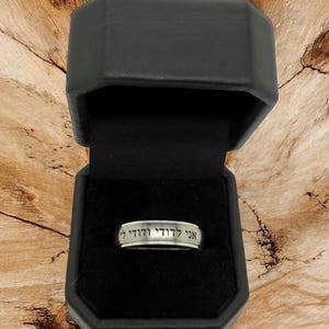 May include: A silver ring with Hebrew text inside a black ring box. The ring is centered in the box, and the text is clearly visible. The box is open, revealing the ring against a black velvet interior.