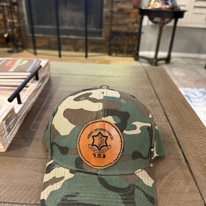 May include: Camouflage baseball cap with a leather patch that reads "Israel Defense Forces" in English and Hebrew. The cap features a brown, green, and tan pattern.