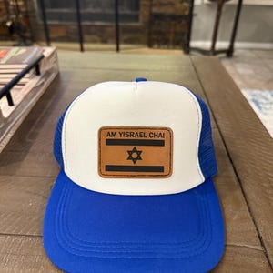 May include: A blue and white trucker hat with a blue brim. The hat features a brown leather patch with the text "AM YISRAEL CHAI" and a Star of David symbol.
