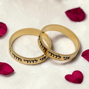 May include: Two gold-toned metal rings featuring a smooth, polished finish. Each ring is engraved with the Hebrew text "אני לדודי ודודי לי" in a clear, black sans-serif font. The rings are positioned overlapping each other on a textured white surface.