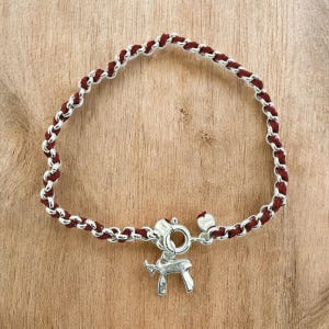 May include: A silver-toned bracelet with a burgundy woven chain design. The bracelet features a small silver charm in the shape of an animal and a clasp closure. The bracelet is displayed on a wooden surface.
