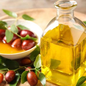 Puede incluir: Una botella de vidrio transparente llena de un líquido amarillo dorado, probablemente aceite, se encuentra sobre una superficie de madera. La botella tiene una boca ancha y un tapón de corcho. Bayas rojas y hojas verdes están esparcidas alrededor de la botella.