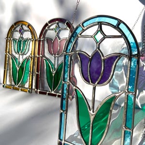 May include: Three stained glass panels depicting tulip flowers in shades of purple, pink, and yellow. Each panel has a silver-colored metal frame and is suspended by a chain. The tulip petals are colored with green leaves.