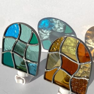 May include: Two stained glass night lights. One features shades of blue and green, the other has amber, orange, and brown hues. Each has a silver-colored metal frame and a white plug.