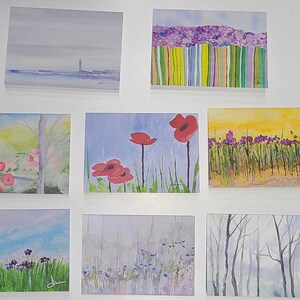 May include: A collection of eight watercolour paintings on display. The paintings feature various subjects, including a seascape, a field of flowers, and a forest scene.