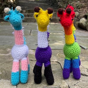 Puede incluir: Tres peluches de jirafa de crochet, cada una con un esquema de color diferente que representa una bandera del orgullo. La primera jirafa es azul, rosa y blanca. La segunda jirafa es amarilla, blanca, morada y negra. La tercera jirafa es roja, naranja, amarilla, verde, azul y morada.
