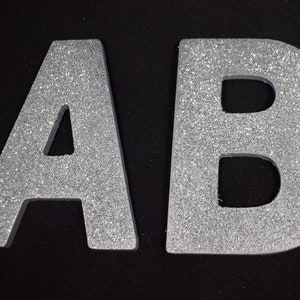 May include: Three silver glitter letters, A, B, and C, are shown against a black backdrop. The letters are in a block style and appear to be made of a solid material, with a sparkly, textured surface. The image is well-lit, highlighting the glitter.