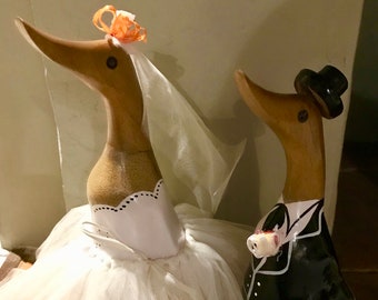 May include: A wooden duck figurine dressed as a bride with a white tulle skirt and a veil, standing next to a wooden duck figurine dressed as a groom in a black tuxedo.