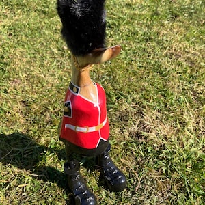 Könnte beinhalten: Holzfigur eines Enten in der Uniform eines britischen Wachmanns. Die Ente trägt einen roten Mantel mit goldenen Verzierungen, einen schwarzen Hut mit Pelzbesatz und schwarze Stiefel.