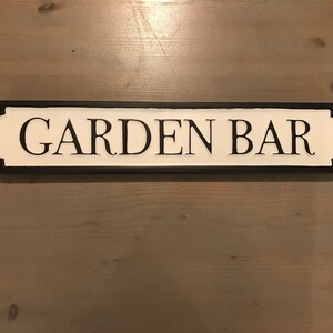 May include: Black and white sign with the text "GARDEN BAR" in black lettering on a white background. The sign has a black border and rounded edges.