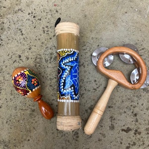 Puede incluir: Un conjunto de tres instrumentos musicales: una maraca de madera con patrones coloridos, un tubo de bambú con un diseño de tortuga azul y un pandero de mano de madera con platillos de plata.