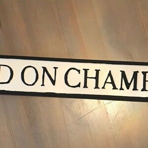 May include: A white sign with black lettering that reads "RAISED ON CHAMPAGNE". The sign is made of wood and has a black border.