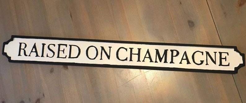 May include: A white sign with black lettering that reads "RAISED ON CHAMPAGNE". The sign is made of wood and has a black border.