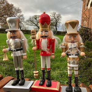 Peut inclure: Trois figurines de casse-noisette en bois, chacune avec un design et un schéma de couleurs différents. La première est argentée avec une barbe blanche et un chapeau noir, la deuxième est rouge avec une couronne dorée et une barbe blanche, et la troisième est dorée avec une barbe blanche et un chapeau noir. Les trois sont debout sur des socles en bois.