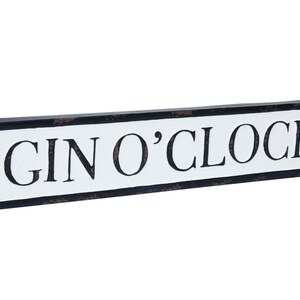 May include: A white sign with black lettering that reads "GIN O'CLOCK" on a black background. The sign is shaped like a street sign and has a distressed look.
