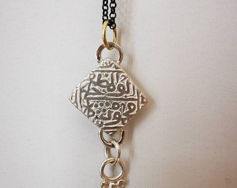 May include: A silver pendant necklace with a square-shaped charm featuring intricate Arabic calligraphy. The charm is attached to a black chain with a brass clasp. The pendant hangs from a silver chain with a small decorative bead.