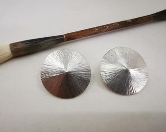 May include: Two silver-colored, textured, circular earrings with a radial pattern are displayed. A wooden-handled calligraphy brush with a white bristle tip is positioned above the earrings. The earrings have a cone-like shape in the center.