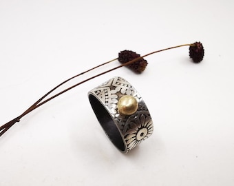 May include: A silver ring with a textured design and a gold bead accent. The ring is on a white background.